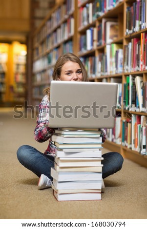 Smiling student sitting on library floor using laptop on pile of books in college
