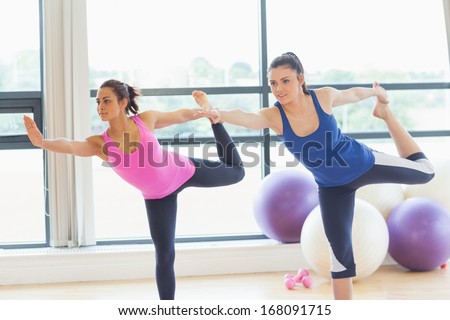 Two fit young women doing the balancing yoga pose in a bright fitness studio