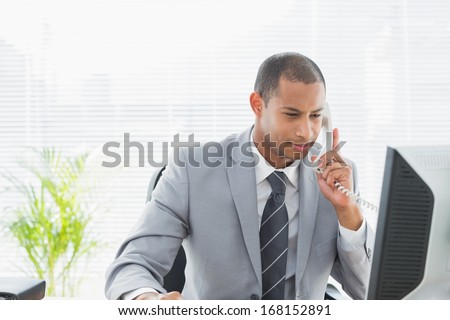 Concentrated young businessman using computer and phone at office desk
