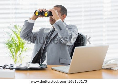Serious young businessman looking through binoculars at office desk