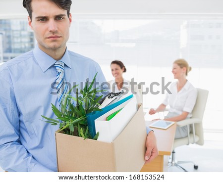Portrait of a young businessman leaving office with his belongings and colleagues in background