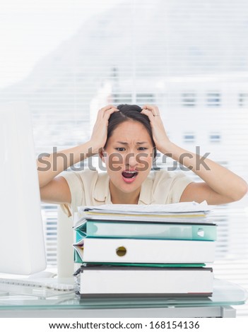 Angry young businesswoman shouting with stack of folders at office desk