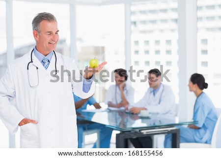 Portrait of a confident doctor holding apple with group around table in background at hospital