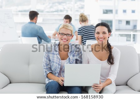 Young women using laptop with colleagues in background at a creative bright office