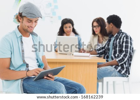 Smiling casual man using digital tablet with group of colleagues behind in a bright office