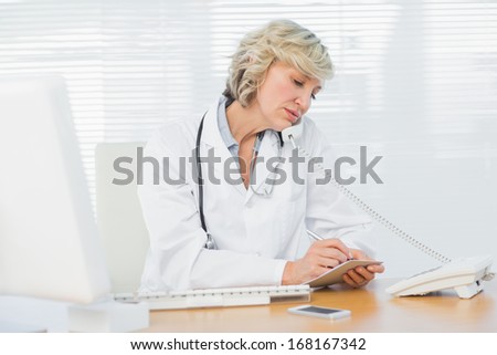 Concentrated female doctor using phone while writing notes by computer at medical office