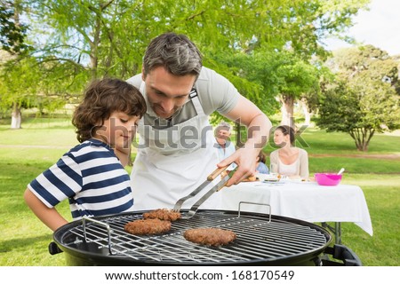 Man And Son Barbecuing With Family In The Background At Park