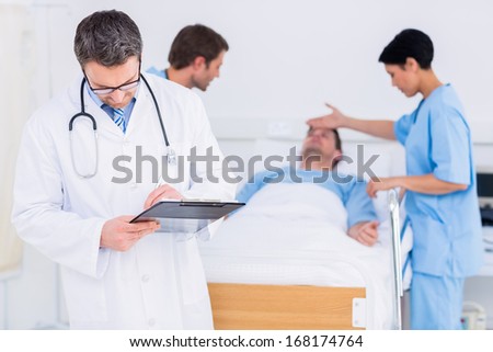 Doctor writing reports with patient and surgeon in background at the hospital