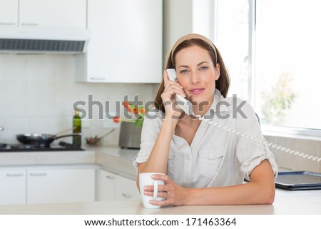 Portrait of a smiling young woman with coffee cup using landline phone in the kitchen at home