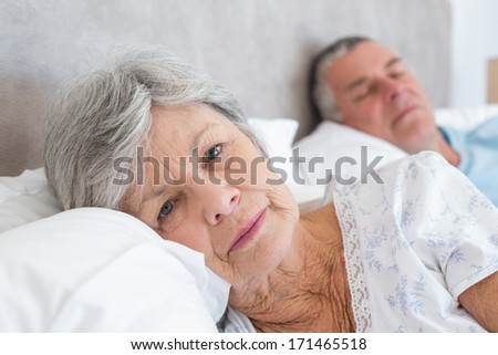 Portrait of sad senior woman lying with man in background at home