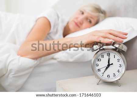 Blurred sleepy mature woman extending hand to alarm clock in bed at home
