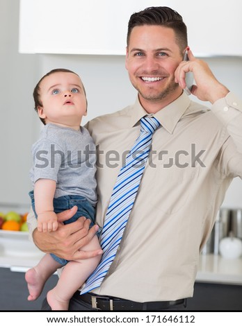 Portrait of handsome businessman using cellphone while carrying baby boy in house