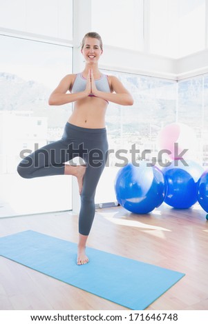 Portrait of a fit smiling young woman standing in tree pose at a bright gym
