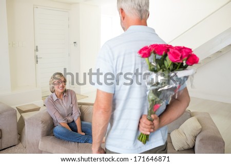 Rear view of a man holding bouquet behind his back with woman sitting on couch at home