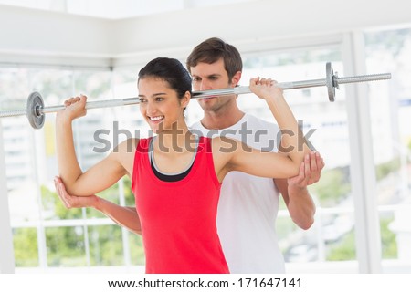 Male trainer helping happy young fit woman to lift the barbell in the gym