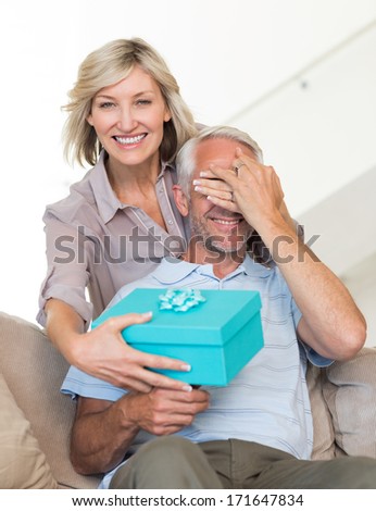 Portrait Of A Smiling Woman Surprising Mature Man With A Gift On Sofa At Home