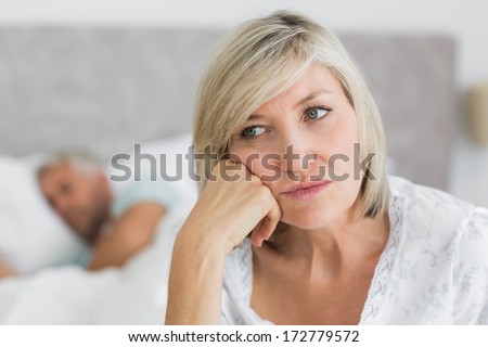 Close-Up Of A Tensed Mature Woman Sitting In Bed With Man In Background At Home