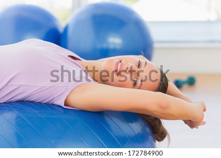 Side view of a smiling fit woman lying on exercise ball at a bright gym