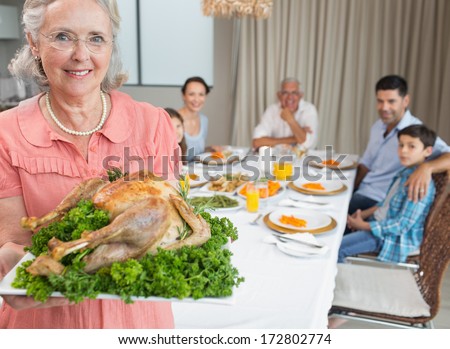 Portrait of a grandmother holding chicken roast with family at dining table in the house