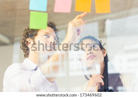 Businessman And Businesswoman Reading Adhesive Notes On Glass Wall In Office
