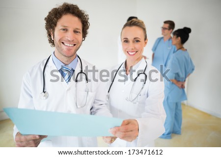 Portrait of happy doctors holding file with colleagues in background at hospital