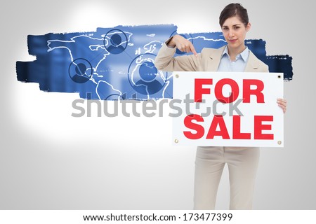 Estate agent holding and pointing to for sale sign against city scene in a room