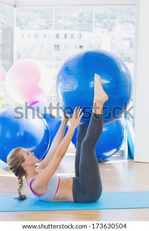 Side view of a sporty young woman holding exercise ball between legs in fitness studio