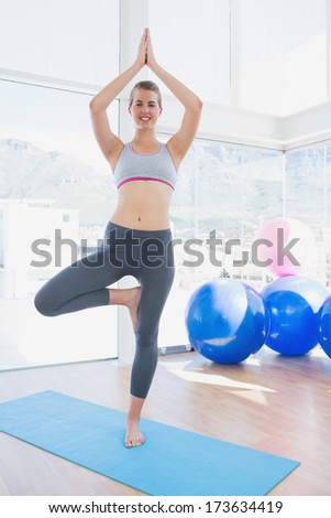 Portrait of a fit smiling young woman standing in tree pose at a bright gym