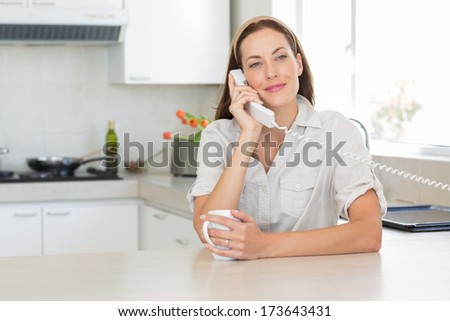 Portrait of a smiling young woman with coffee cup using landline phone in the kitchen at home