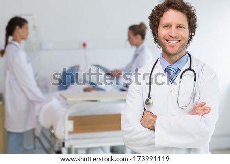 Portrait of male doctor standing arms crossed with colleagues examining patient in hospital