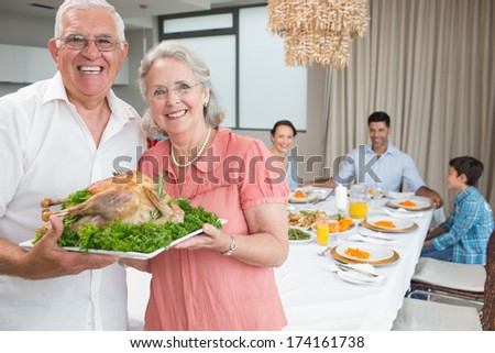 Portrait of grandparents holding chicken roast with family at dining table in the house