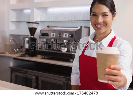 Happy young barista offering cup of coffee to go smiling at camera in a cafe