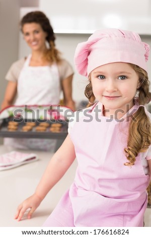 Cute girl wearing pink apron and chefs hat smiling at camera at home in kitchen