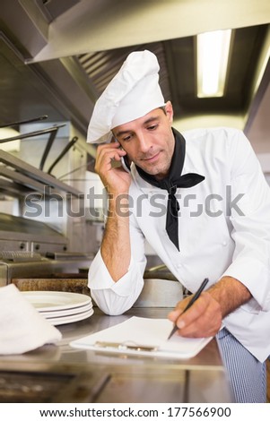 Concentrated male cook writing on clipboard while using cellphone in the kitchen