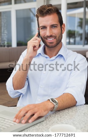 Smiling businessman talking on phone using his laptop in patio of restaurant