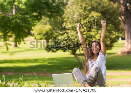 Portrait of a cheerful young woman raising hands with laptop in the park