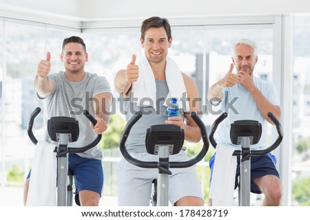 Portrait of men on exercise bikes gesturing thumbs up at gym