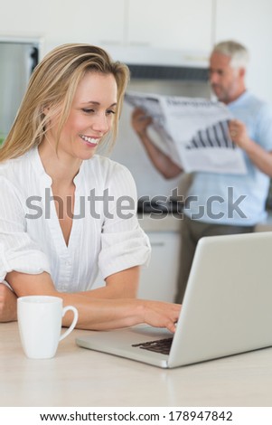 Smiling woman using laptop with partner standing with the paper at home in the kitchen