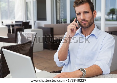 Frowning businessman talking on phone using his laptop in patio of restaurant