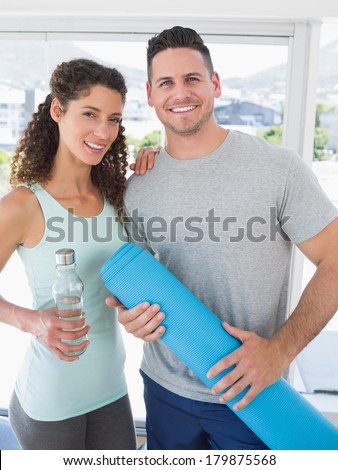 Portrait of happy couple holding water bottle and exercise mat in exercise room