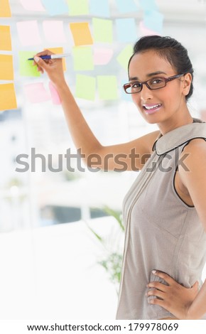 Designer writing on sticky notes on window smiling at camera in creative office