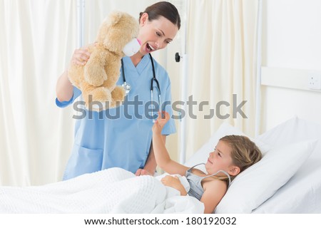 Playful female doctor entertaining sick girl with teddy bear in hospital bed