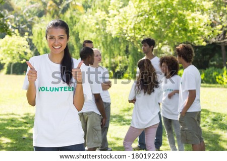 Portrait of young female volunteer gesturing thumbs up at park with friends in backgorund