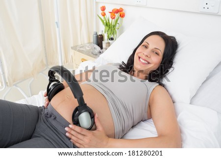 Portrait of happy pregnant woman with headphones on her belly lying in bed at hospital