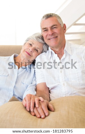 Retired couple holding hands on couch smiling at camera at home in living room
