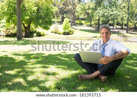 Full length portrait of mature businessman with laptop sitting on grass in park