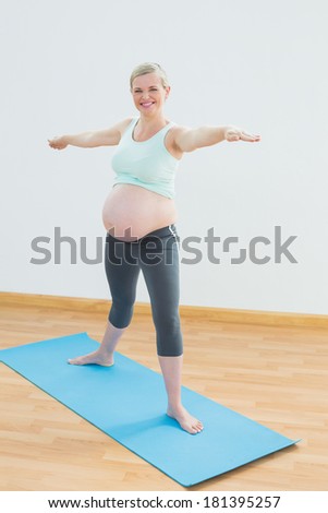 Blonde pregnant woman doing yoga on blue mat smiling at camera in a fitness studio