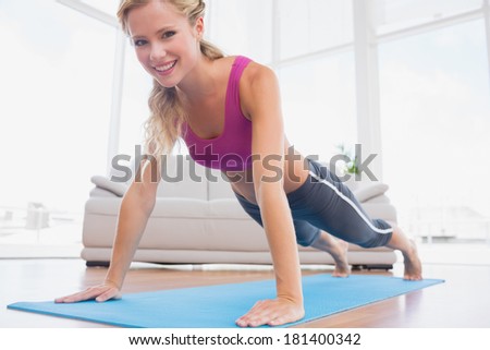 Strong blonde in plank position on exercise mat smiling at camera at home in the living room