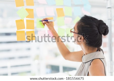 Smiling designer writing on sticky notes on window in creative office
