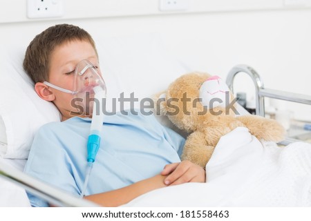 Sick boy wearing oxygen mask sleeping beside teddy bear in hospital bed